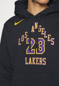Hoodie noir avec logo Nike jaune. Présente des textes violets et jaunes : "LOS ANGELES", "23" et "LAKERS" dans différentes polices. Le matériau est doux et texturé.