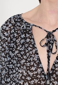 Blouse noire transparente avec petit imprimé floral bleu et blanc, avec un décolleté en V profond noué au niveau de la clavicule avec des liens en tissu assortis.