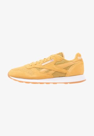 Zapatilla Reebok de ante amarillo y malla con suela blanca y suela exterior de goma, con el logo de Reebok y la bandera del Reino Unido en el lateral.