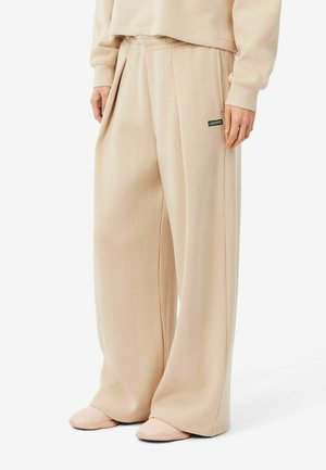 Pantalon beige à jambes larges avec plis et poches latérales, petit logo Lacoste vert sur la poche droite, porté avec un haut beige assorti et des chaussures.