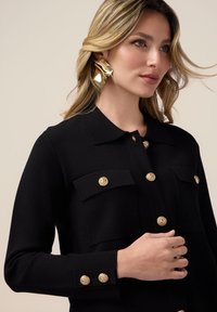 Giacca camicia nera a maglia con colletto, due tasche sul petto e dettagli in bottone dorato su tasche e polsini. Texture liscia.