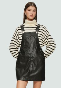 Zwarte leren overall jurk met verstelbare bandjes, voorzak, gedragen over een wit met zwarte gestreepte coltrui. Gladde textuur.