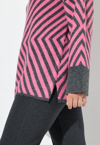 Rosa und grauer gestreifter Pullover mit einem Zickzackmuster, aus weichem Material gefertigt, mit abgerundetem Saum und gerippten Bündchen, kombiniert mit dunklen Leggings.