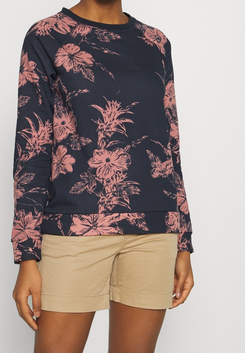 Personne portant un sweat-shirt bleu marine avec de grands motifs floraux roses et un short beige, debout devant un fond clair uni.