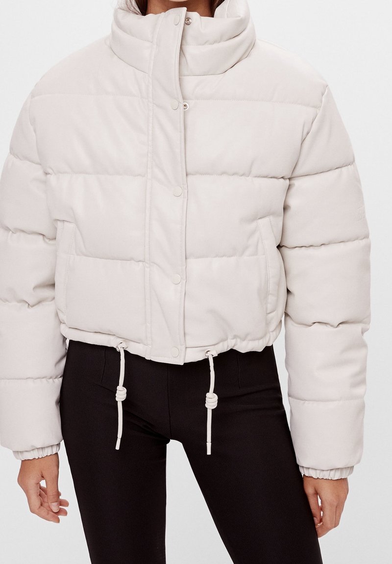 Femme portant une doudoune beige clair avec boutons-pression et cordons à l'ourlet, associée à un pantalon noir ajusté.