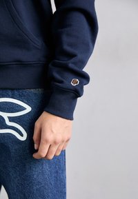Navy sweatshirt with a soft texture, featuring a small brown ice cream cone patch on the cuff, paired with denim featuring a white outline design.