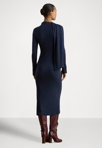 Tamsiai mėlyna ilgarankovė suknelė aptempto silueto su drapiruotu nugaros apdaru, derinama su bordo odiniais kelio aukščio batais su kulnu ir sagtimi.