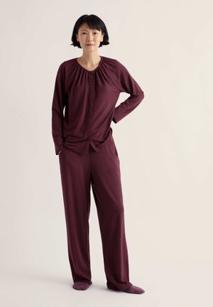 Femme debout, une main sur la hanche, portant un haut bordeaux à manches longues boutonné assorti à un pantalon ample avec des chaussettes, sur fond uni.