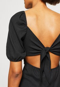 Robe noire avec des manches bouffantes et un dos ouvert noué. Texture côtelée avec une taille élastique.