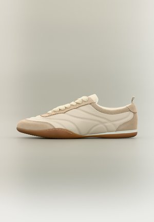 Baskets beige et blanches profil bas avec détails en suède, lacets blancs et semelle en caoutchouc marron sur fond neutre.