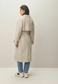 Lys beige trenchcoat med bælte i taljen, struktureret krave og lukket manchet, båret over blå jeans og hvide sneakers.