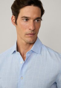 Camisa de botones azul claro con un patrón de cuadrícula, hecha de una tela texturizada, que presenta un cuello clásico y botones de plástico blanco.