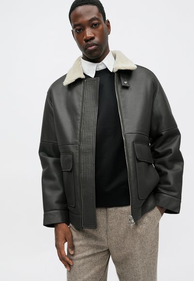 Tmavá kožená bunda s jasným umelým kožušinovým golierom, so zipsom a prešívanými detailmi na prednej strane, v kombinácii s čiernym sveterom a vzorovanými nohavicami.