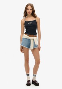Schwarzes Camisole mit weißem "SUPERDRY NYC" Text, Jeansshorts mit ausgefranstem Saum, leichter Pullover in der Taille gebunden, gestreifte Socken und dunkle Loafers.