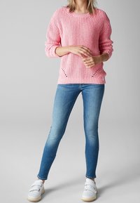 Różowy sweter dzianinowy z teksturowanym wzorem, zestawiony z dopasowanymi niebieskimi dżinsami i białymi trampkami z rzepami.