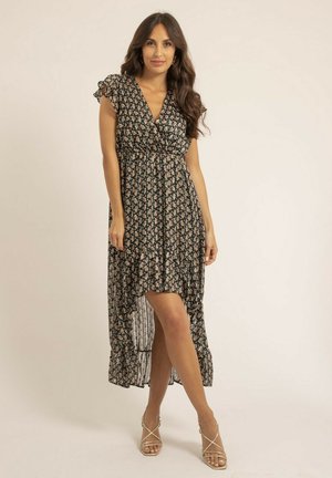 Femme debout portant une robe noire à fleurs courte devant et longue derrière avec des manches à volants et des talons beige à fines lanières, sur un fond uni.