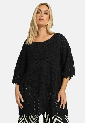 Poncho negro de punto con un patrón texturizado, mangas anchas y bordes en forma de concha, combinado con pantalones negros y blancos con patrón.