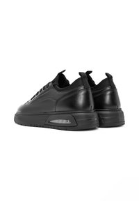 Las zapatillas de cuero negras cuentan con un acabado suave, punta redonda, diseño con cordones y un cojín de aire visible en el talón, con una suela de goma texturizada.