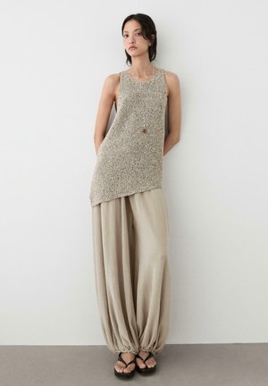 Kobieta stojąca przy ścianie, ubrana w bezrękawnikową plecioną tunikę, luźne beżowe spodnie ściągnięte przy kostkach, sandały i długi naszyjnik z zawieszką.