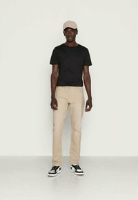 T-shirt preto, jeans bege e ténis pretos com solas brancas. Usando um boné rosa claro, de pé contra um fundo simples.