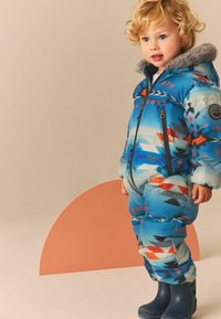 Un traje de nieve acolchado de un brillante color azul con patrones geométricos en rojo y negro, capucha con borde de piel sintética, cierre frontal y acompañado de botas de goma azules.