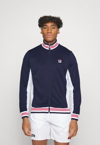 Chaqueta navy con cremallera, paneles laterales blancos y cuello a rayas rojas y blancas. Presenta un pequeño logo en el lado izquierdo del pecho y puños acanalados.