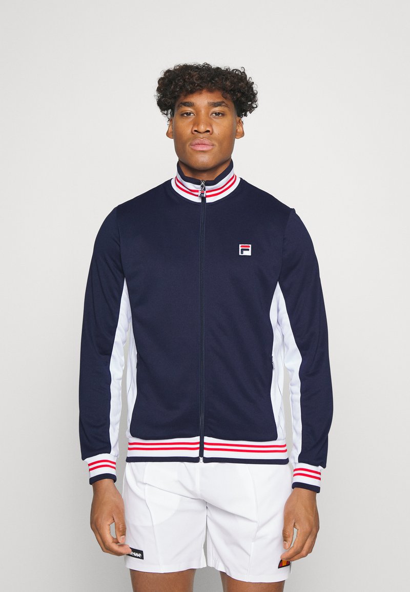 Chaqueta navy con cremallera, paneles laterales blancos y cuello a rayas rojas y blancas. Presenta un pequeño logo en el lado izquierdo del pecho y puños acanalados.