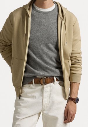 Mann trägt eine beige Zip-up-Kapuze, grauen Pullover über weißem Hemd, weiße Hose mit braunem Gürtel und braune Armbanduhr, steht vor weißem Hintergrund.