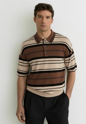 Mand iført en brun, beige og sort polo med korte ærmer og vandrette striber, samt sorte bukser, stående mod en hvid væg.