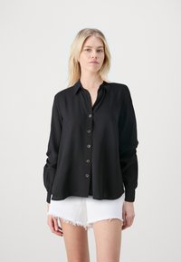 Camisa negra de manga larga, con botones, cuello y corte relajado. Presenta un tejido suave, cierre frontal con botones y dobladillo dividido. Combinada con pantalones cortos blancos.