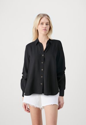 Schwarzes Langarm-Hemd mit Knopfleiste, Kragen und lockerer Passform. Aus weichem Stoff, mit Vorderknopfverschluss und gespaltenem Saum. Kombiniert mit weißen Shorts.