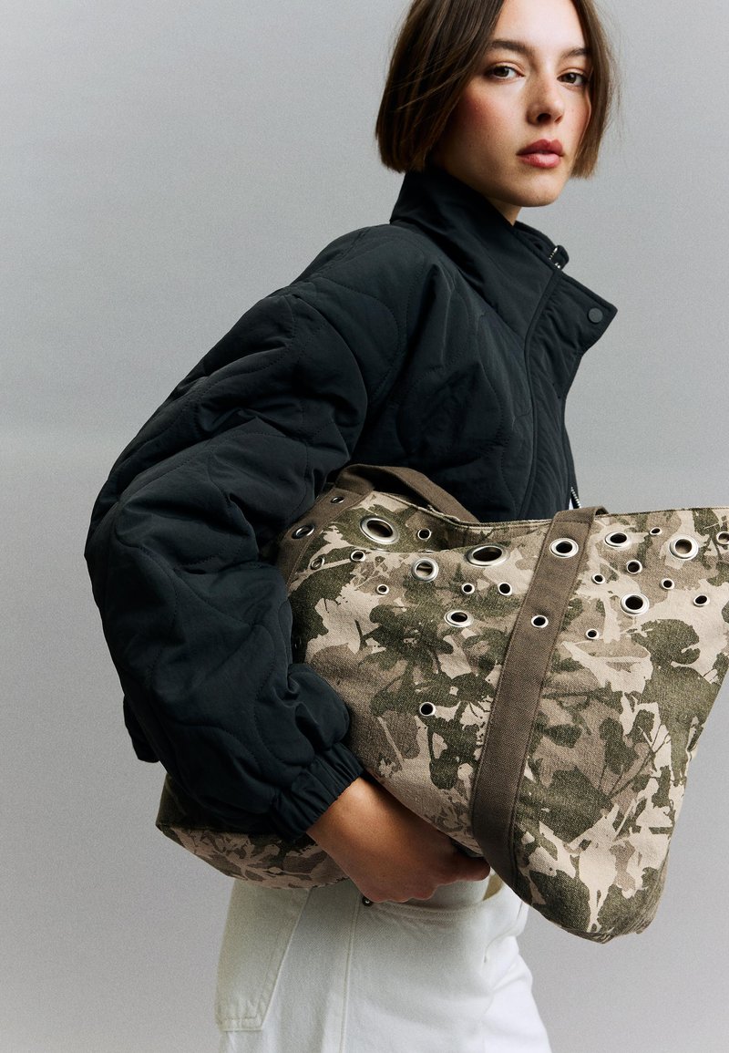 Ung person iført en sort quiltet jakke holder en stor beige og grøn floral lærredstaske med metaløjer.