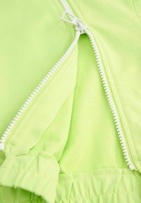 Zielona, zapinana na zamek odzież o gładkiej fakturze, z białym zamkiem błyskawicznym i elastycznymi mankietami. Materiał wydaje się miękki i wygodny.