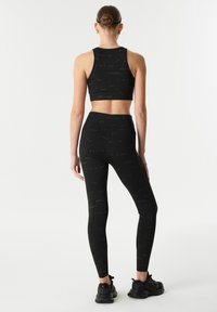Černý sportovní crop top a legíny s opakujícím se textovým vzorem. Hladký povrch, přiléhavý design, doplněný o černé sportovní boty.