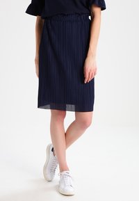 Granatowa spódnica plisowana noszona z czarną bluzką i białymi trampkami. Modelka stoi na jasnym tle, eksponując szczegóły stroju.