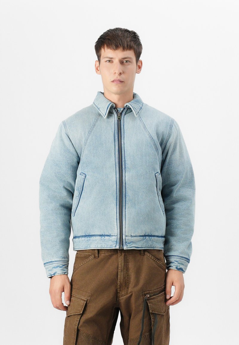 Veste en jean bleu clair avec des manches raglan, fermeture éclair à l'avant, deux poches latérales et coutures contrastées en bleu foncé.