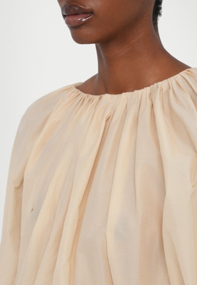 Helle beige Bluse mit einem gerafften, runden Halsausschnitt; aus weichem, fließendem Material mit subtiler Textur und Drapierungsdetails.