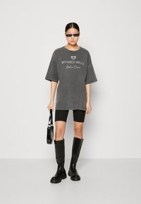 Szürke, oversized póló, rajta fehér "BEVERLY HILLS" felirattal, fekete biciklisnadrággal és térd fölé érő fekete csizmával párosítva. Fekete kézitáskát tart.