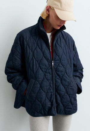 Personne portant une veste matelassée foncée avec les mains dans les poches, une casquette beige, un pantalon clair et la tête légèrement penchée vers le bas sur un fond uni.