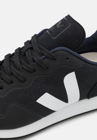 Black athletic shoe featuring a textured upper, white logo accent, and a rubber sole with a sleek, modern design. Laces are dark gray.