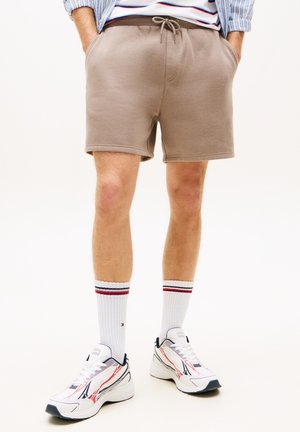 Mann trägt beige Shorts mit Kordelzug, weiße Sportschuhe mit roten und schwarzen Details, weiße Sportsocken mit roten und schwarzen Streifen, steht.