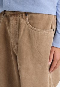 Brown corduroy trousers with vertical ribbing, featuring a front pocket, black button accents, and a relaxed fit. Light blue shirt in background.