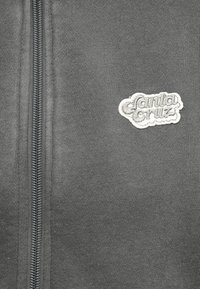 Γκρι πλεκτό ύφασμα με κάθετο μαύρο φερμουάρ στα αριστερά και λευκή κεντημένη ετικέτα με το λογότυπο "Santa Cruz" στα δεξιά.