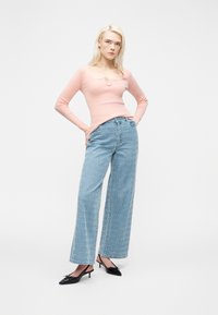 Γυναίκα με ξανθά μαλλιά που φοράει ροζ ανοιχτό ρούχο με μακριά μανίκια, ψηλόμεσα φαρδιά παντελόνια με σχέδια και μαύρα πέδιλα με τακούνι και λουράκι στο πίσω μέρος.