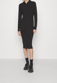 Schwarzes Rippstrickkleid mit kragenähnlichem Ausschnitt, langen Ärmeln und figurbetontem Design, kombiniert mit schwarzen Schnürschuhen und Crew-Socken.