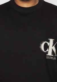 Svart sweatshirt med en vit spraymålad "ck"-logotyp och varumärkestext på nedre högra sidan. Tyget är mjukt och har rundad halsringning.