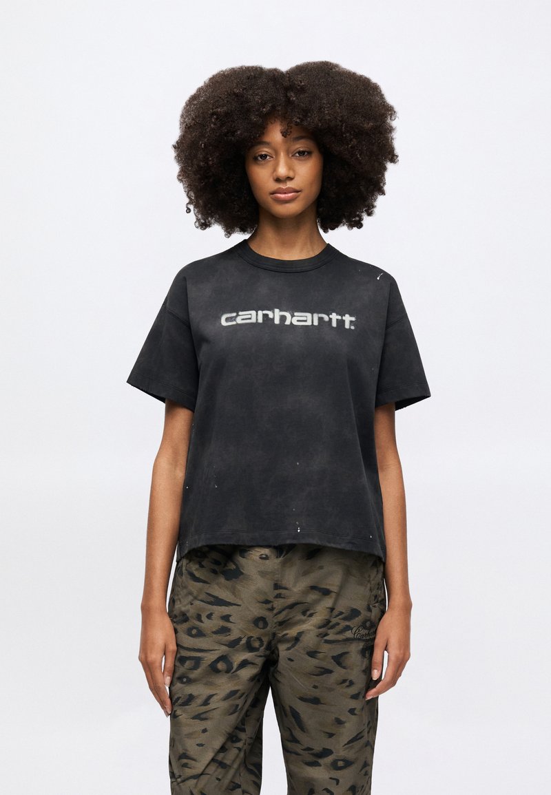 Sort t-shirt med korte ærmer og en let cropped pasform. Har et gråt "carhartt"-logo på forsiden. Kombineret med mønstrede olivengrønne bukser.