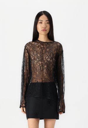 Haut noir à manches longues en dentelle florale transparente, doté d'un panneau arrière noir uni, porté avec une jupe noire ; motif floral complexe sur l'ensemble.