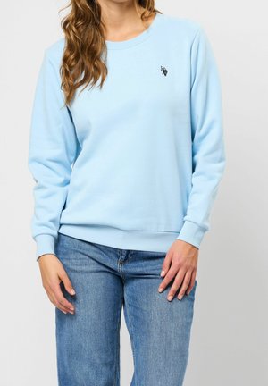 Vrouw draagt een lichtblauwe sweater met lange mouwen en een klein zwart logo op de borst, gecombineerd met een blauwe spijkerbroek, staand tegen een effen grijze achtergrond.