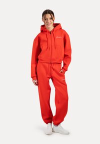 Sweat-shirt à capuche rouge et pantalon de jogging assorti en matériau doux, avec une poche avant, un cordon de serrage et un logo brodé. Baskets blanches.